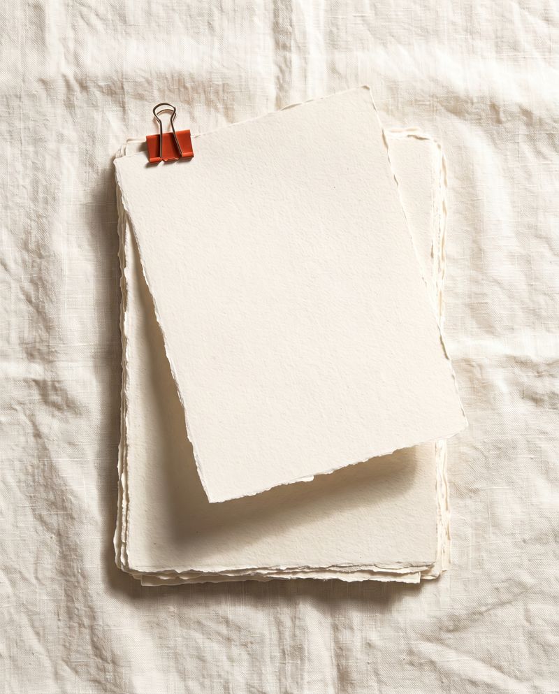 A neat stack of blank deckled sheets on linen, held at one corner by a small vermilion clip